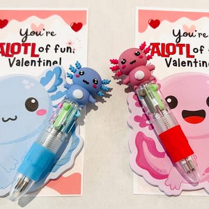 K&ouml;nnte beinhalten: Zwei Valentinstagskarten mit Axolotl-Illustrationen und mehrfarbigen Stiften. Auf den Karten steht "You're ALOTL of fun, Valentine!". Ein Axolotl ist blau, das andere rosa. Die Stifte haben mehrere Tintenfarben.