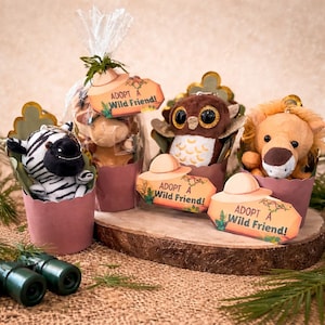 May include: A collection of plush animal toys, including a zebra, giraffe, owl, and lion, presented in decorative paper cups. Each toy has a tag that reads "ADOPT A Wild Friend!" The arrangement is set on a wooden surface.