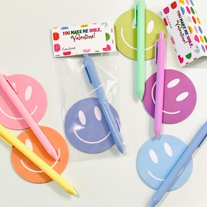 May include: A collection of colorful pens and smiley face-shaped notepads. The pens are in shades of pink, yellow, green, and blue. The notepads have a smiley face design and the text "YOU MAKE ME SMILE, Valentine! From friend."