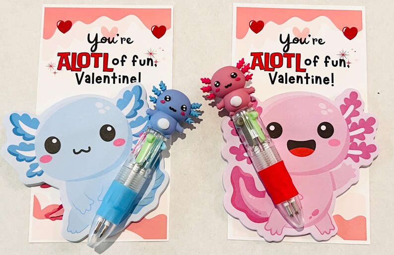 K&ouml;nnte beinhalten: Zwei Valentinstagskarten mit Axolotl-Stiften. Eine Karte zeigt einen blauen Axolotl und Stift, die andere einen rosa Axolotl und Stift. Jede Karte tr&auml;gt den Text "You're ALOT of fun, Valentine!" und Herzdekorationen.