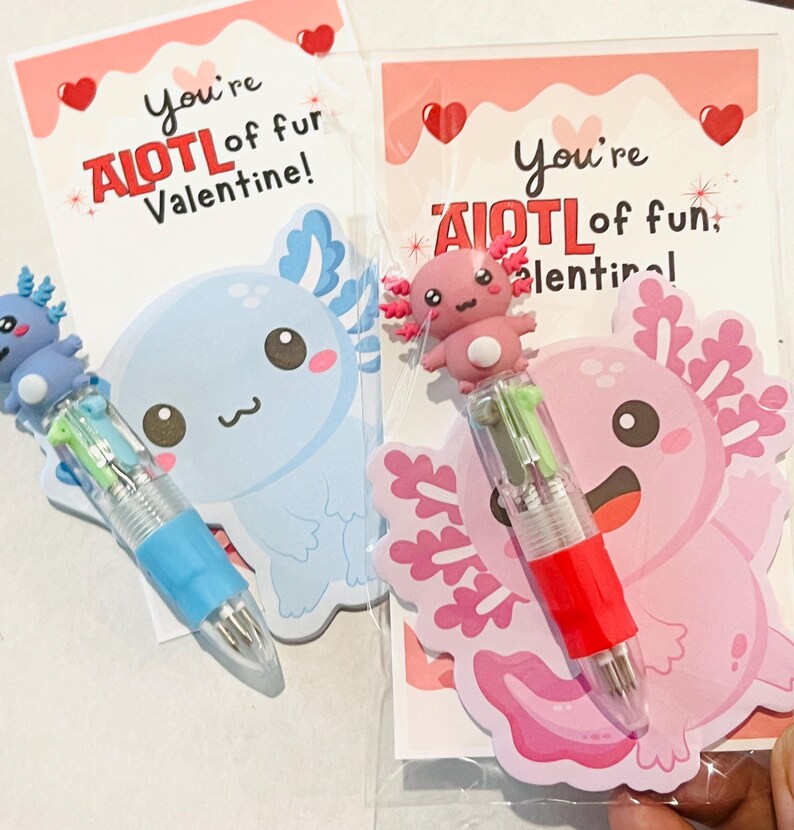 K&ouml;nnte beinhalten: Zwei Valentinstagskarten mit Stiften zum Thema Axolotl. Eine Karte zeigt einen blauen Axolotl und den Text "You're AIOTL of fur Valentine!" Die andere Karte hat einen rosa Axolotl und den Text "You're AIOTL of fun, Valentine!"