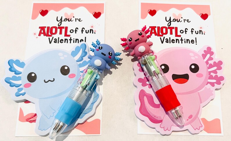 K&ouml;nnte beinhalten: Zwei Valentinstagskarten mit Axolotl-Designs. Eine Karte zeigt einen blauen Axolotl mit einem blauen Stift, die andere einen rosa Axolotl mit einem roten Stift. Jede Karte tr&auml;gt den Text "You're ALOTL of fun, Valentine!"