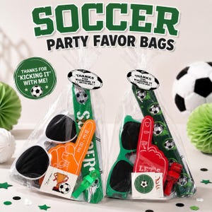 May include: Two clear plastic party favor bags filled with soccer-themed items. Each bag contains sunglasses, a foam finger, a whistle, and a green inflatable cheering stick. The bags are sealed with a tag that reads, Thanks for kicking it with me.