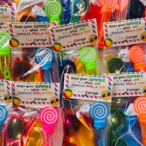 May include: A set of colorful summer party favor bags with a variety of items, including sunglasses, bubble wands, and small inflatable balls. Each bag has a tag that says "Hope your summer is a splash with bubbles, ball, & sun!" and is signed "From: Carsyn".
