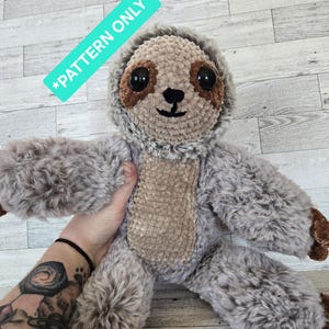 May include: A plush sloth toy made of textured gray and brown yarn with large black eyes and a stitched nose. A teal rectangular tag in the upper left corner displays the white text *PATTERN ONLY*. The sloth has a light brown belly.