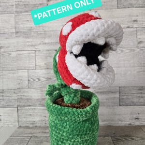 May include: Crocheted Piranha Plant from the Super Mario Bros. video game series. The plant has a red head with white spots and a white mouth, green stem and leaves, and is potted in a green pot. The text "*PATTERN ONLY*" is on the image.