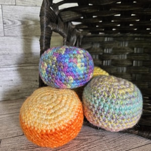 May include: Four colorful crocheted balls in shades of orange, yellow, blue, purple, and green are displayed. The balls are in front of a dark brown woven basket. The balls are handmade and have a textured appearance.