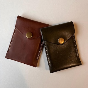 May include: Two leather wallets, one brown and one black, with a brass button closure. Both wallets have a flap pocket on the front.