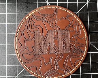 May include: A brown leather coaster with a stitched edge and a topographic design. The letters "MI" are embossed in the center.