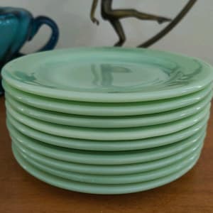 Puede incluir: Una pila de ocho platillos redondos de vidrio en color verde menta claro. Cada platillo presenta un acabado liso y brillante con un borde ligeramente elevado y un centro poco profundo. El vidrio tiene una calidad semitranslúcida, característica de la cristalería de jadeíta, con variaciones sutiles en el color verde pálido a lo largo de toda la pila.