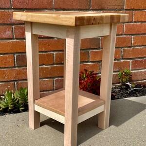 May include: A small wooden end table with a light wood finish. The table has a square top and a shelf underneath. The table is sitting on a concrete surface.