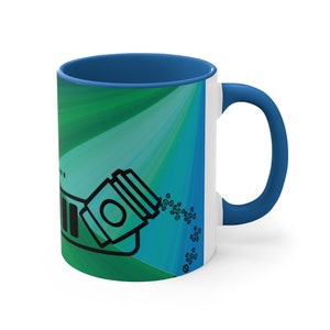 May include: A white ceramic coffee mug with a blue handle and a green and blue abstract design. The mug features a black and white illustration of a coffee cup spilling coffee beans.