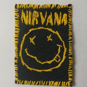 May include: A black rectangular patch featuring yellow embroidered text and a smiley face design. The word "NIRVANA" is stitched across the top. The smiley face has crossed-out eyes and a tongue sticking out. The edges are also stitched in yellow.