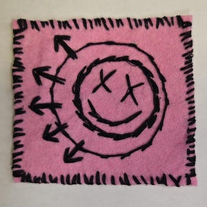 Peut inclure: Un écusson carré en feutrine rose avec une broderie noire faite à la main. Le motif représente un visage souriant circulaire avec des yeux en forme de X et une bouche courbée, entouré de cinq flèches pointant vers l'extérieur. Les bords sont finis au point de surjet noir.
