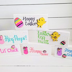 May include: Six wooden blocks with Easter-themed decorations and sayings. The blocks are white with a speckled finish. The sayings include "Happy Easter", "Hey Peeps!", "Easter Egg Hunt", "Lil' Chick", "Hunny Bunny", and "Hoppy Easter Peep".
