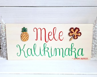 Op de afbeelding: Een houten bord met de tekst "Mele Kalikimaka - Jimmy Buffett" geschreven in groene en rode verf. Het bord is versierd met een gele ananas en een rode hibiscusbloem.