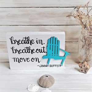 May include: Wooden sign with the quote "breathe in. breathe out. move on." attributed to Jimmy Buffett. The sign features a teal blue cutout of a beach chair.
