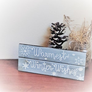 May include: A rustic wooden sign with a distressed blue paint finish. The sign has two lines of white text that reads "Warmest Winter Wishes" and is decorated with white snowflake designs.