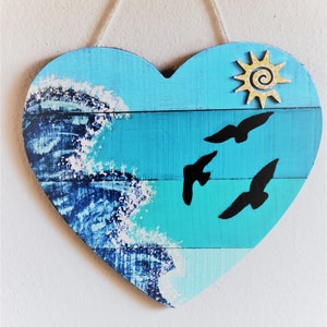 May include: A blue and white heart-shaped wooden sign with a painted ocean wave design and silhouettes of birds flying. A yellow sun with a spiral design is in the upper right corner.