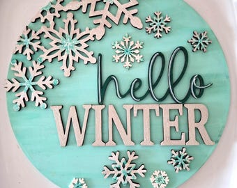 May include: A round, turquoise wooden sign with the words "hello WINTER" in white and green lettering. The sign is decorated with various sizes of silver and turquoise snowflakes. The sign is placed on a white surface.