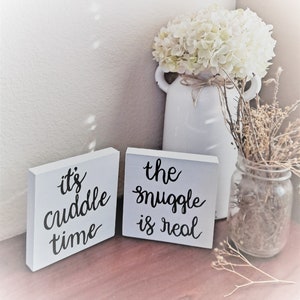 May include: Two white wooden signs with black calligraphy. The first sign says "it's cuddle time". The second sign says "the snuggle is real".