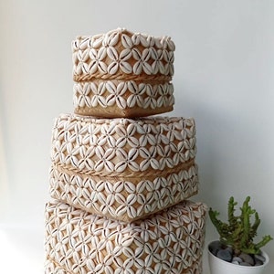 May include: Three square woven baskets made of natural materials, decorated with white seashells in a floral pattern. The baskets are stacked on top of each other, with the smallest on top.