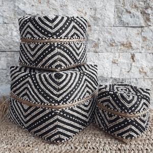 Puede incluir: Un conjunto de tres cajas de almacenamiento decorativas con tapas. Las cajas están adornadas con un patrón geométrico de cuentas negras y blancas. Una cuerda de fibra natural rodea cada caja. Las cajas están dispuestas sobre una estera tejida.