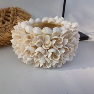 May include: A white seashell bowl with a round shape. The bowl is made of many small seashells that are arranged in a circular pattern. The bowl is ideal for holding small items, such as jewellery or keys.