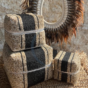 Puede incluir: Tres cajas cuadradas hechas de cuentas tejidas en beige y negro. Las cajas están apiladas una encima de la otra, con la caja más grande en la parte inferior y la caja más pequeña en la parte superior. Las cajas están sentadas en una estera tejida.