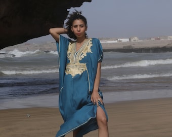 May include: A woman wearing a teal blue kaftan with gold embroidery stands on a sandy beach. The kaftan has a V-neckline and long sleeves. The woman is wearing a gold bracelet on her wrist. The text "Elegance Marrakech by Maryamah" is visible at the bottom of the image.