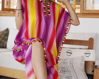 May include: A woman wearing a colorful striped kaftan with tassels. The kaftan is a vibrant mix of pink, yellow, red, and orange stripes. The woman is sitting on a chair with a white cushion and a Moroccan-style rug.