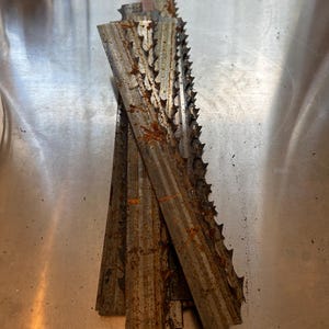 May include: Close-up of several weathered metal blades with jagged edges. The blades are stacked, showing signs of rust and wear. The metal has a dull, aged appearance. The blades are likely for a tool.