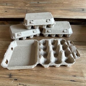 May include: Several empty, light brown egg cartons are displayed on a wooden surface. Three closed cartons are stacked, while one is open, revealing the individual egg compartments. The cartons are made of a recycled paper material.