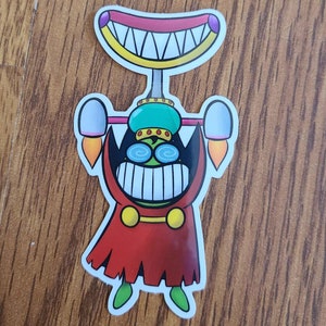 May include: A cartoon sticker of a green and red creature with a large toothy grin. The creature is wearing a green crown and a red robe. The sticker is on a wooden surface.
