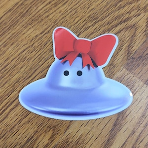May include: A blue and purple flying saucer with a red bow on top. The saucer has two black eyes.