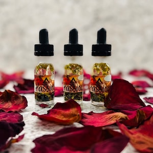 May include: Three small glass bottles with dropper tops filled with a clear liquid and dried flowers. The bottles have a black label with a triangle and the word "Passion" on them. The bottles are sitting on a white surface with red rose petals.