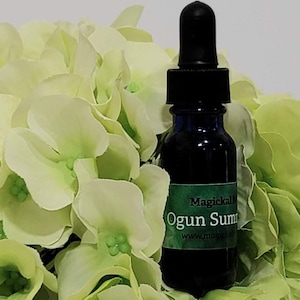 Puede incluir: Una pequeña botella de vidrio azul con un gotero negro en la parte superior. La botella está llena de un líquido oscuro y tiene una etiqueta verde que dice "Magickal Ogun Summit" y "www.magickal". La botella está sentada sobre un lecho de flores blancas y verdes.