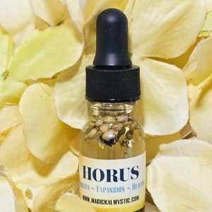 May include: A small glass bottle with a black dropper top filled with a clear liquid and small, light brown seeds. The bottle has a white label with the text "HORUS" in black lettering and the words "ABILITY ~ EXPANSION ~ HEALING" in smaller black lettering. The text "WWW.MAGICKALMYSTIC.COM" is printed in black at the bottom of the label.