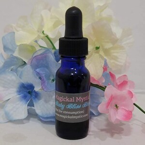 Op de afbeelding: Een donkerblauwe glazen fles met een zwarte druppelaar, gevuld met een donkere vloeistof. Het etiket luidt "Magickal Mystic Baby Bliss Bloo" en "Not for consumption". De fles is omgeven door kunstbloemen in wit, blauw en roze.