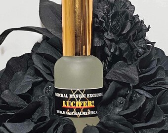 May include: A clear glass bottle with a gold cap and a black label that reads "Magickal Mystic Exclusive Lucifer! www.magickalmystic.ca". The bottle is surrounded by black fabric flowers.