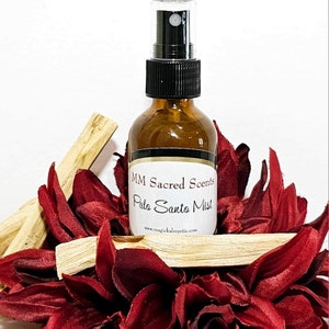Op de afbeelding: Een bruine glazen fles met een zwarte spuitdop en een wit etiket met de tekst "MM Sacred Scents Palo Santo Mist" en "www.magicalmystic.com". De fles is omringd door stukken Palo Santo hout.