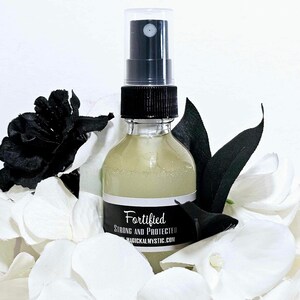 Puede incluir: Una botella de vidrio transparente con un pulverizador negro y una etiqueta blanca que dice "Fortified Strong and Protected magicalmystic.com". La botella está rodeada de pétalos de flores blancas y negras.