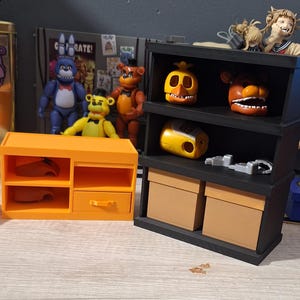 May include: An orange miniature cabinet with two shelves and a drawer, next to a black shelf unit with two tan boxes. The shelves display character heads and a yellow pencil sharpener. Several character figurines are in the background.