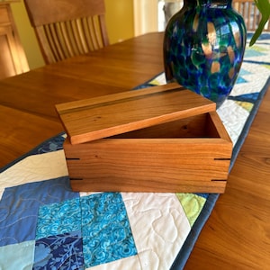 May include: A wooden box with a lid, crafted from light brown wood. The box is open, revealing its interior. The lid is slightly ajar, and the box is placed on a blue and white patterned table runner.