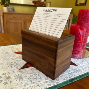 May include: A wooden recipe box with a card that reads "RECIPE" is displayed on a table. The box is a dark brown color and has a slot for the recipe cards. Two red candles are in the background, and the table has a festive runner.