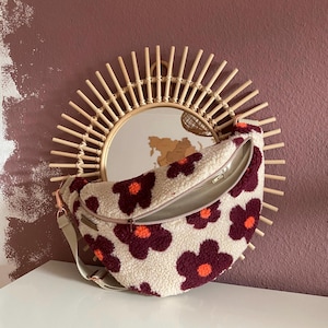 Könnte beinhalten: Cremefarbene Bauchtasche mit einem Blumenmuster in Burgunderrot und Orange. Die Tasche hat einen Reißverschluss und einen beigen Riemen. Die Tasche wird vor einem runden Spiegel mit Sonnenstrahlenrahmen präsentiert.