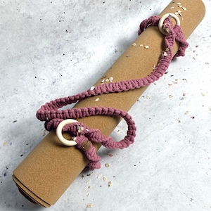 Könnte beinhalten: Eine aufgerollte Kork-Yogamatte mit einem rosafarbenen gehäkelten Riemen und Holzringen. Der Riemen ist um die Matte gebunden und hat ein kleines braunes Etikett mit Text.