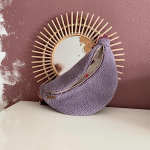 Könnte beinhalten: Eine flauschige, lavendelfarbene Bauchtasche mit Reißverschluss. Die Tasche liegt auf einer weißen Oberfläche vor einem runden Sonnenstrahlspiegel aus Bambus. Der Hintergrund ist in einem gedämpften Malvenfarbton gehalten.