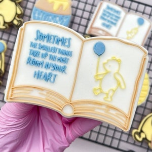 Puede incluir: Una galleta decorada en blanco y amarillo con forma de libro abierto. El libro tiene el texto "Sometimes the smallest things take up the most room in your heart" en la página izquierda y un oso amarillo con un globo azul en la página derecha.