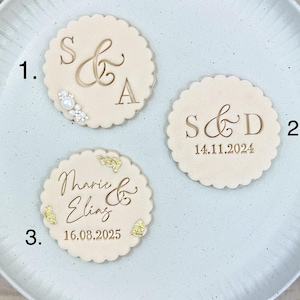 Könnte beinhalten: Drei runde Holzschilder mit personalisiertem Text und dekorativen Akzenten. Das erste Schild sagt "S&A" mit kleinen weißen Perlenakzenten. Das zweite Schild sagt "S&D 14.11.2024". Das dritte Schild sagt "Marie & Elias 16.08.2025" mit goldenen Glitzerakzenten.